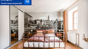 Une chambre avec un cadre de lit en métal, un plancher en bois, une grande fenêtre et une fresque représentant un paysage urbain sur le mur.