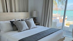 A large bed with white linens and a grey throw blanket, a wooden headboard, a nightstand with a lamp, and a view of the ocean through large windows. - Waterfront Georgetown home, 8ppl,Chef, Boat Tour (Exuma)