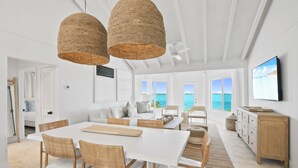 A spacious dining area with a white table and wicker chairs, a white sofa, and a view of the ocean through large windows. - Waterfront Georgetown home, 8ppl,Chef, Boat Tour (Exuma)