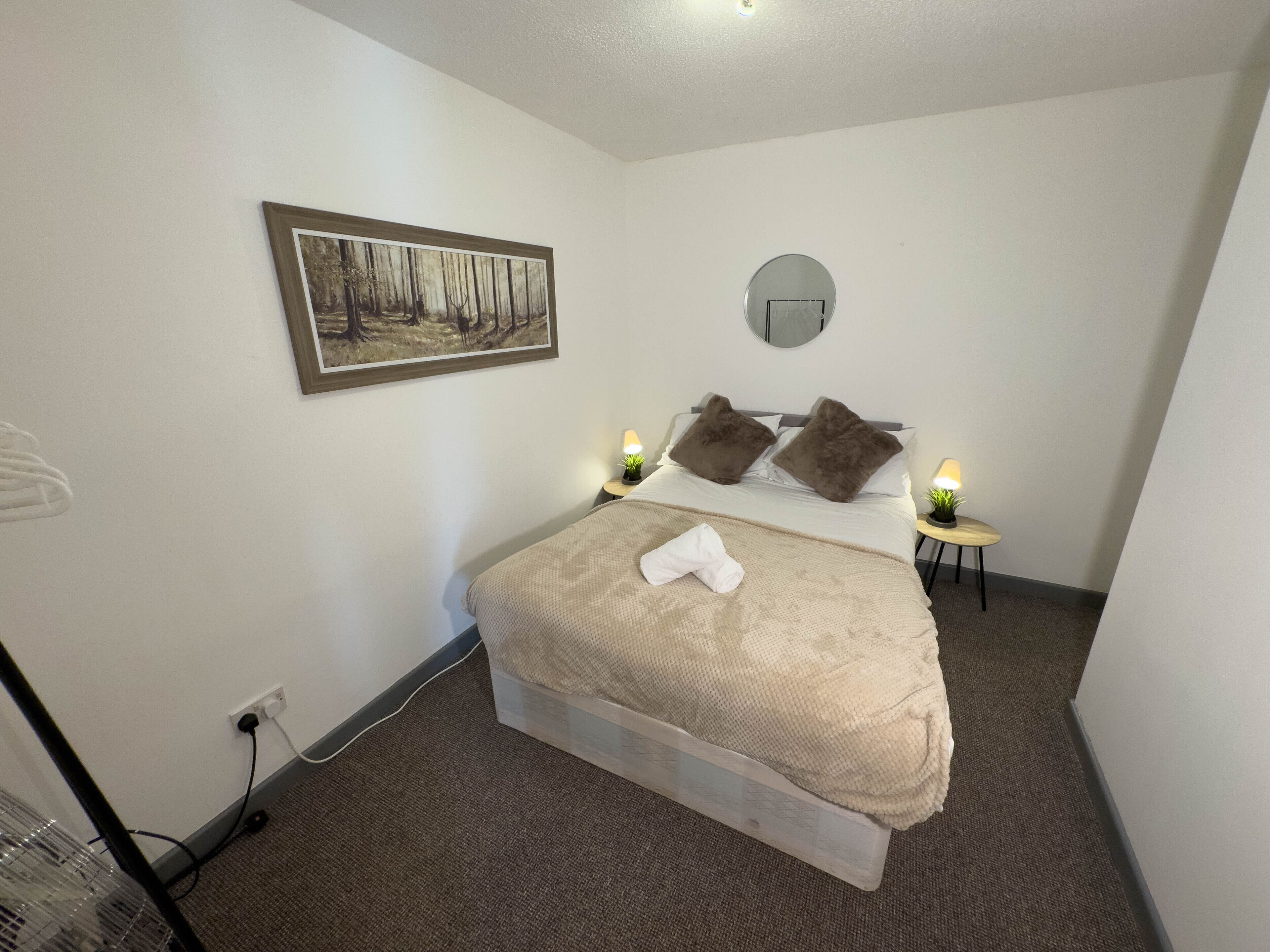 A neatly made bed with a beige blanket and pillows, a small bedside table with a lamp, a wall-mounted mirror, and a framed picture of a forest scene.