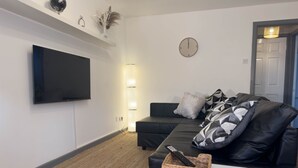 Ein modernes Wohnzimmer mit einem Wand-Fernseher, einem schwarzen Ledersofa mit gemusterten Kissen, einem kleinen weißen Tisch und einem gewebten Korb mit einer Fernbedienung.