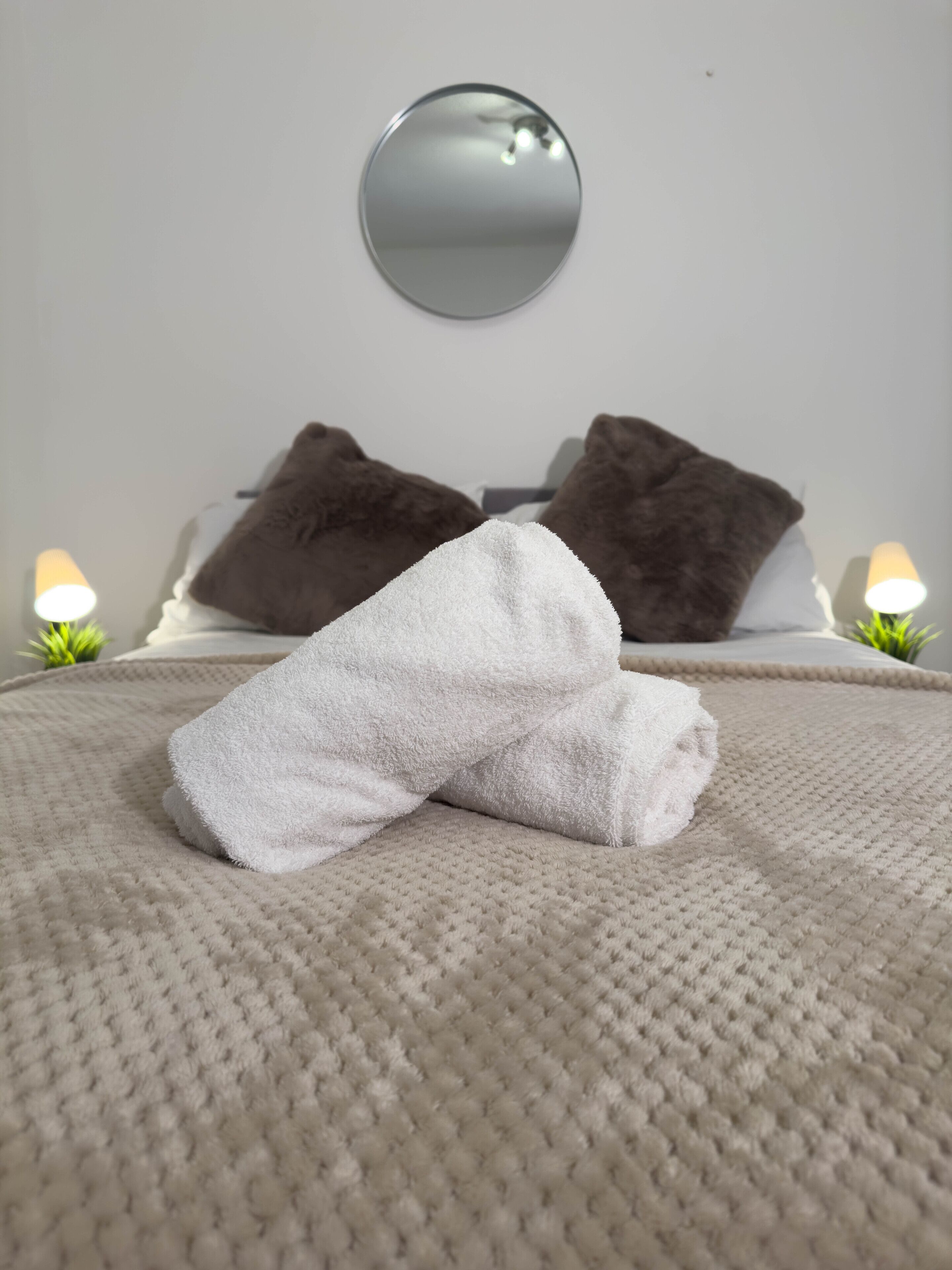 A neatly made bed with a white towel, two brown pillows, and a round mirror on the wall.