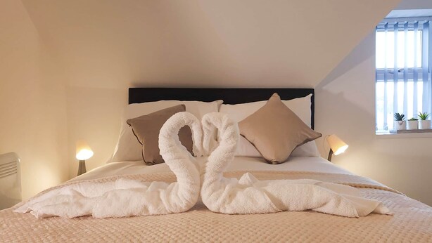 A neatly made bed with a unique towel arrangement, a headboard, bedside lamps, and a window with blinds.