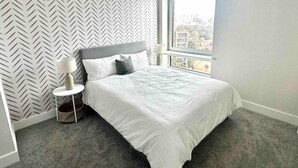 Un dormitorio moderno con cama, mesita de noche, lámpara y un amplio ventanal con vista a la ciudad.