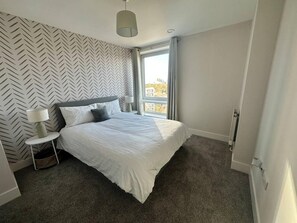 Une chambre moderne avec un grand lit, une table de chevet, une lampe et une fenêtre donnant sur des immeubles.