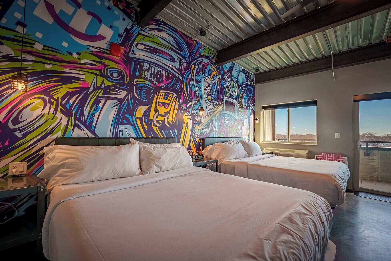 Habitación de hotel con dos camas, un mural abstracto y llamativo en la pared, y un amplio ventanal con vistas al exterior.