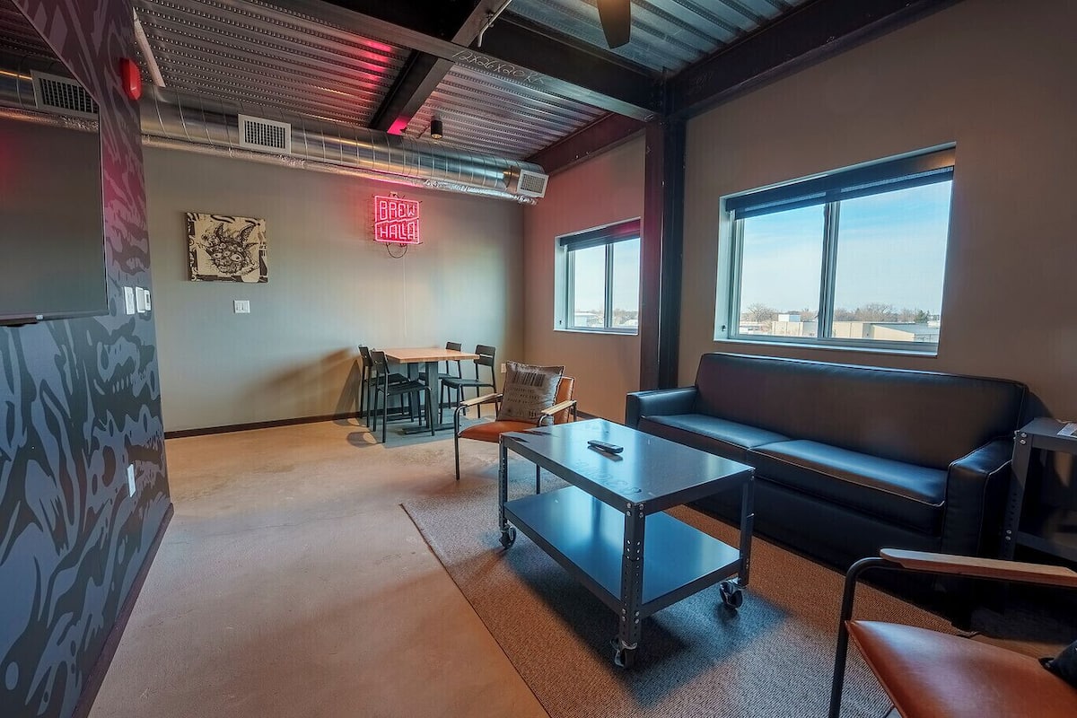 Uma área de estar moderna com sofá, mesa de centro e mesa de jantar com cadeiras. Há um letreiro de neon na parede e grandes janelas que proporcionam iluminação natural.