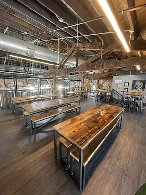 Amplia zona de comedor de estilo industrial con largas mesas de madera y bancos de metal, rodeada de equipos de cocina de acero inoxidable y un techo alto con vigas y conductos a la vista.