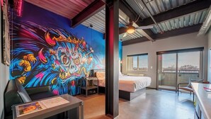 Una habitación de hotel moderna con un mural llamativo, una cama, un escritorio y vistas a través de un ventanal grande.