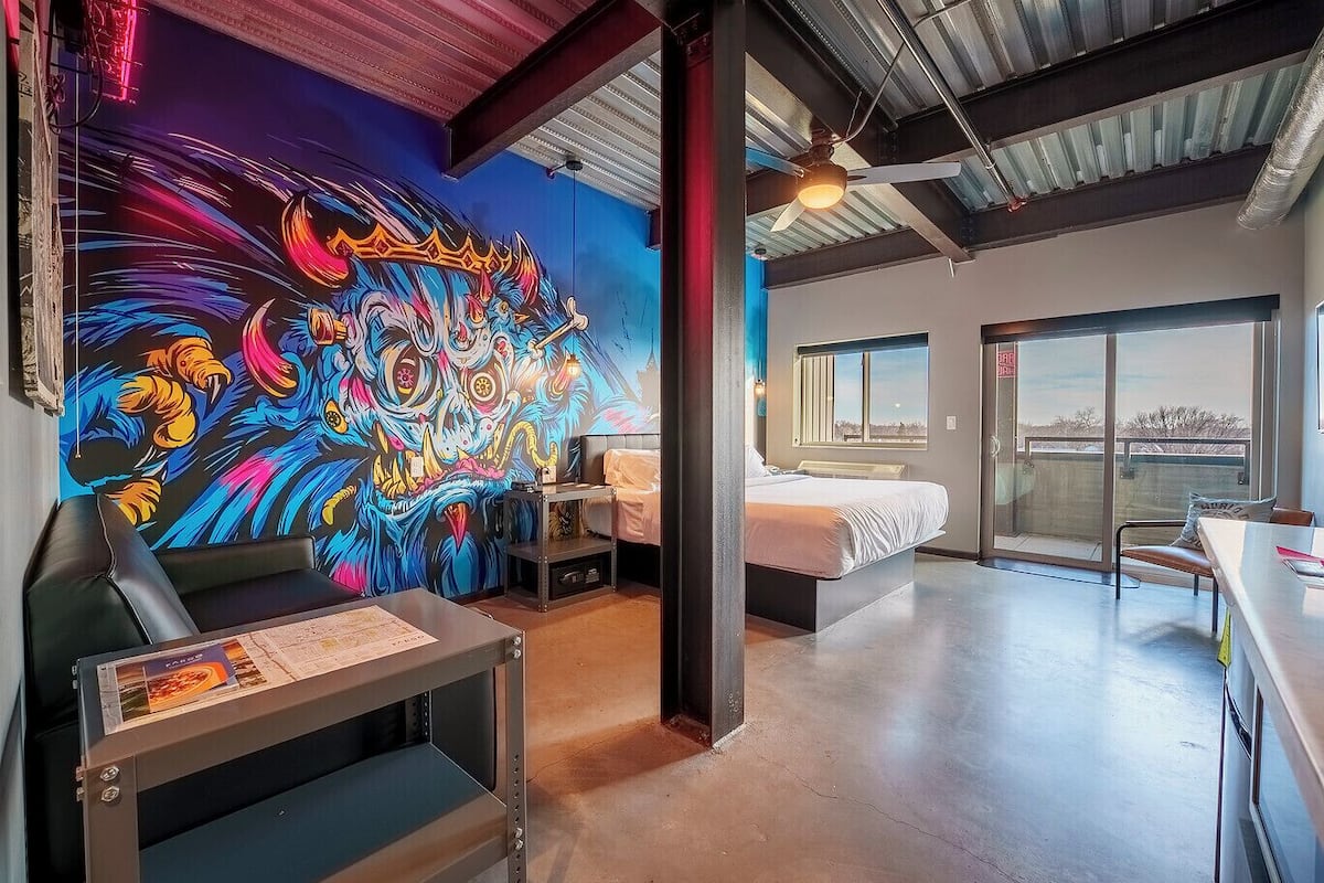 Quarto de hotel moderno com um mural vibrante, uma cama, uma escrivaninha e vista através de uma grande janela.