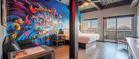 A modern hotel room with a vibrant mural, a bed, a desk, and a view through a large window.