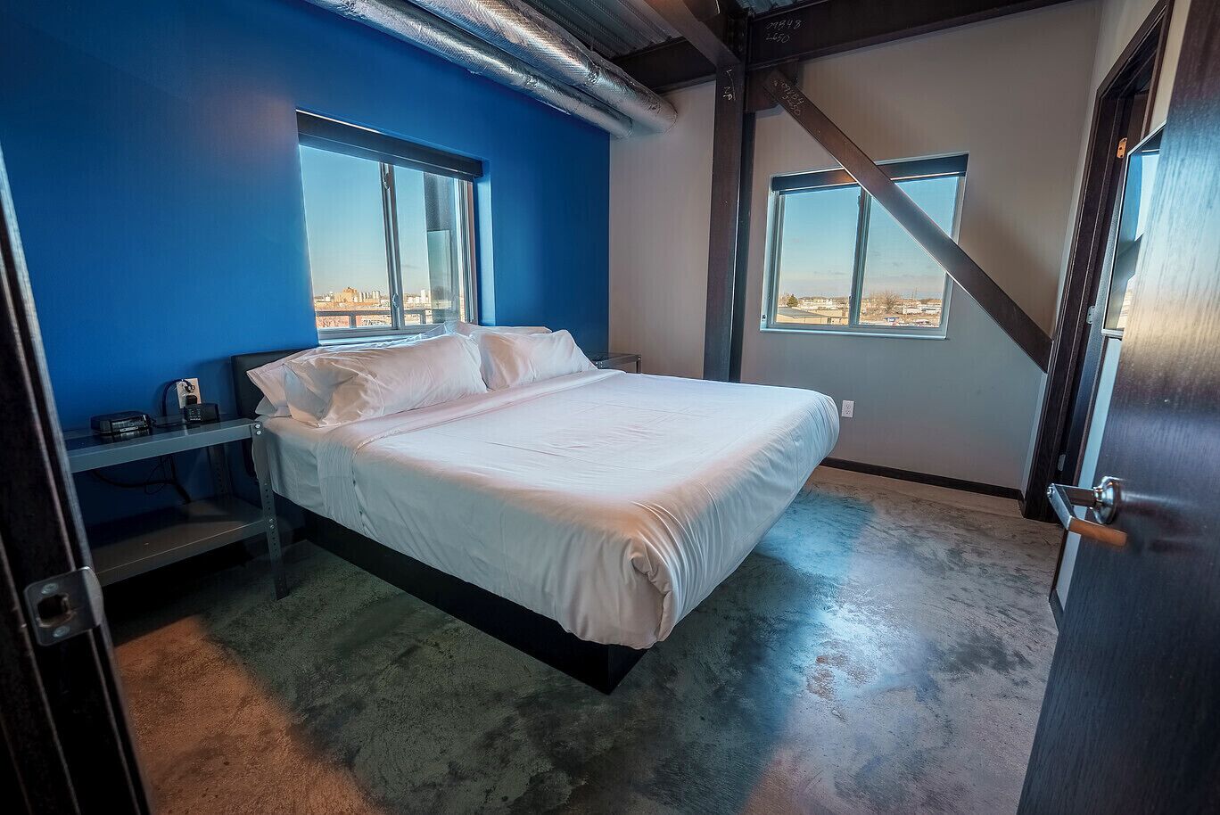 A bedroom with a bed, a nightstand, a desk, and two windows offering a view.