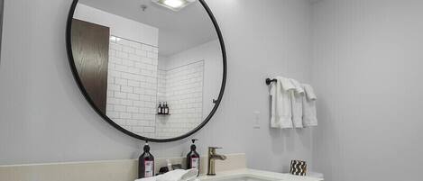 A modern bathroom with a white sink, round mirror, and white toilet. There are towels and toiletries on the counter.