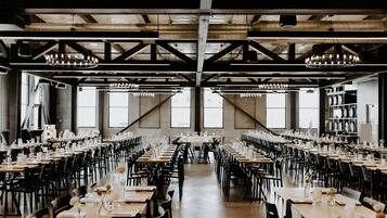 Une vaste salle à manger, dotée de nombreuses tables dressées pour le repas, de grandes fenêtres et d’un plafond haut avec des poutres apparentes.