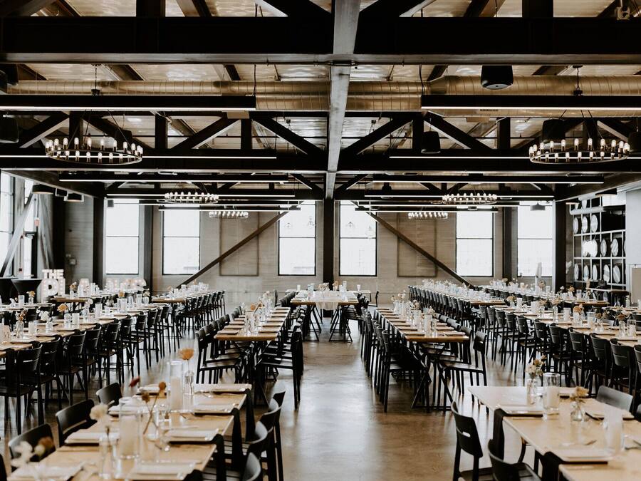 Een ruime eetzaal met meerdere tafels gedekt voor een maaltijd, grote ramen en een hoog plafond met zichtbare balken.