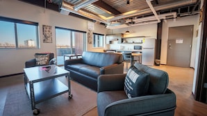 Una sala de estar moderna con un sofá, una mesa de centro y un carrito. Se observa una cocina al fondo con microondas y refrigerador.