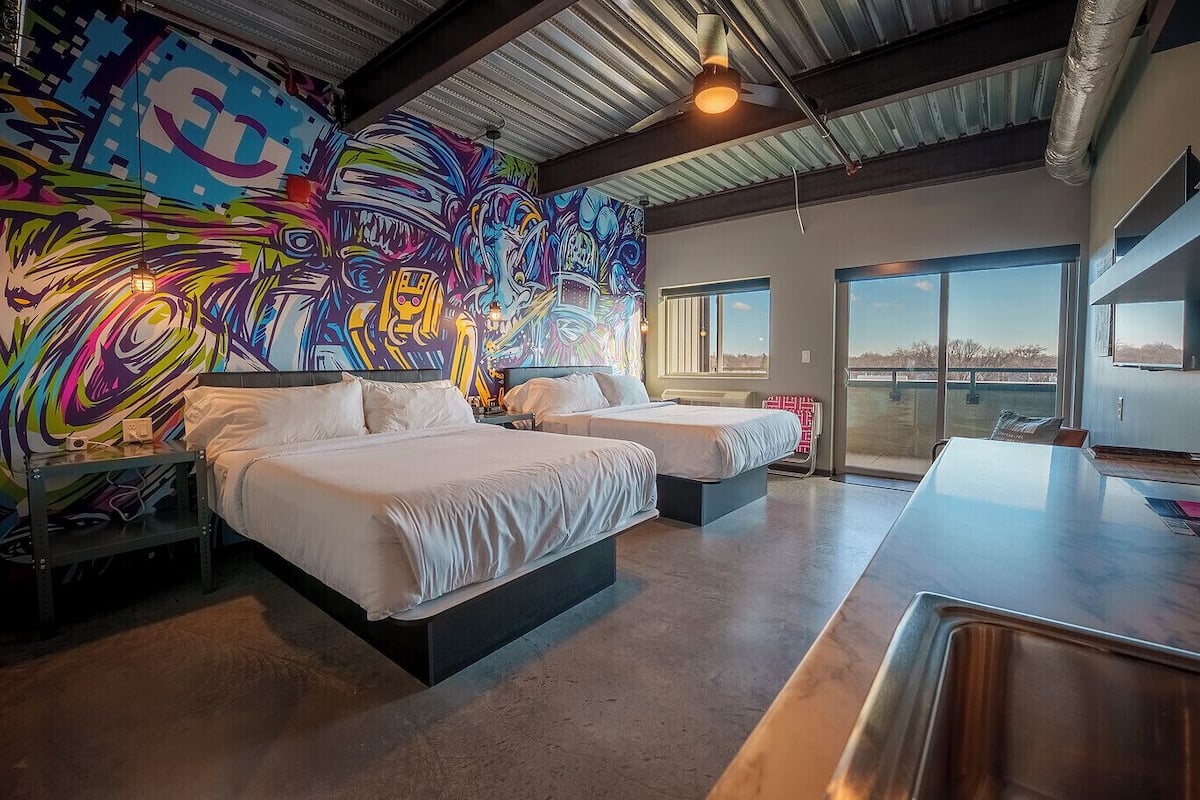 Quarto de hotel moderno com duas camas, um grande mural abstrato e vista para o exterior.