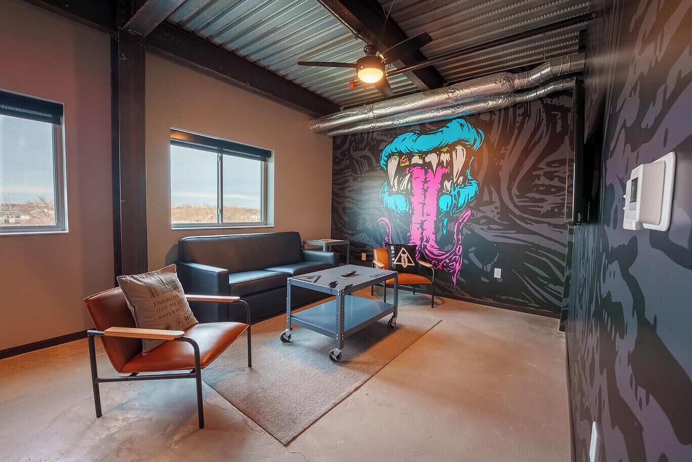 A modern living room with a black leather sofa, a brown leather chair, and a small table. There is a large window and a mural on the wall.