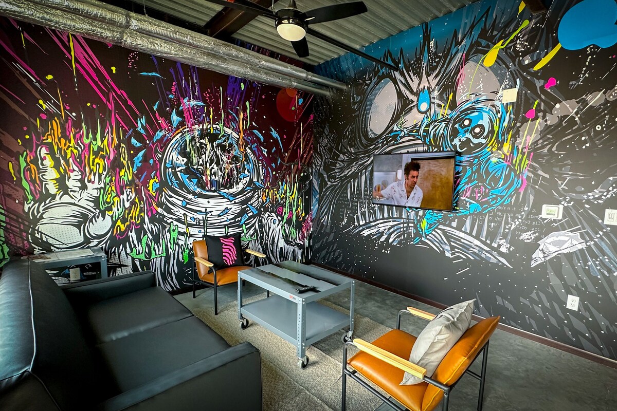 A room with a large abstract mural, a television, and a seating area with a sofa and chairs.