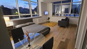 Une chambre moderne avec un lit, une table de chevet et un bureau avec une chaise. De grandes fenêtres offrent une vue sur l’extérieur.