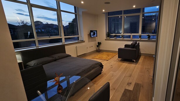 Une chambre moderne avec un grand lit, un bureau et une chaise. De nombreuses fenêtres laissent entrer la lumière naturelle et offrent une vue sur l’extérieur.
