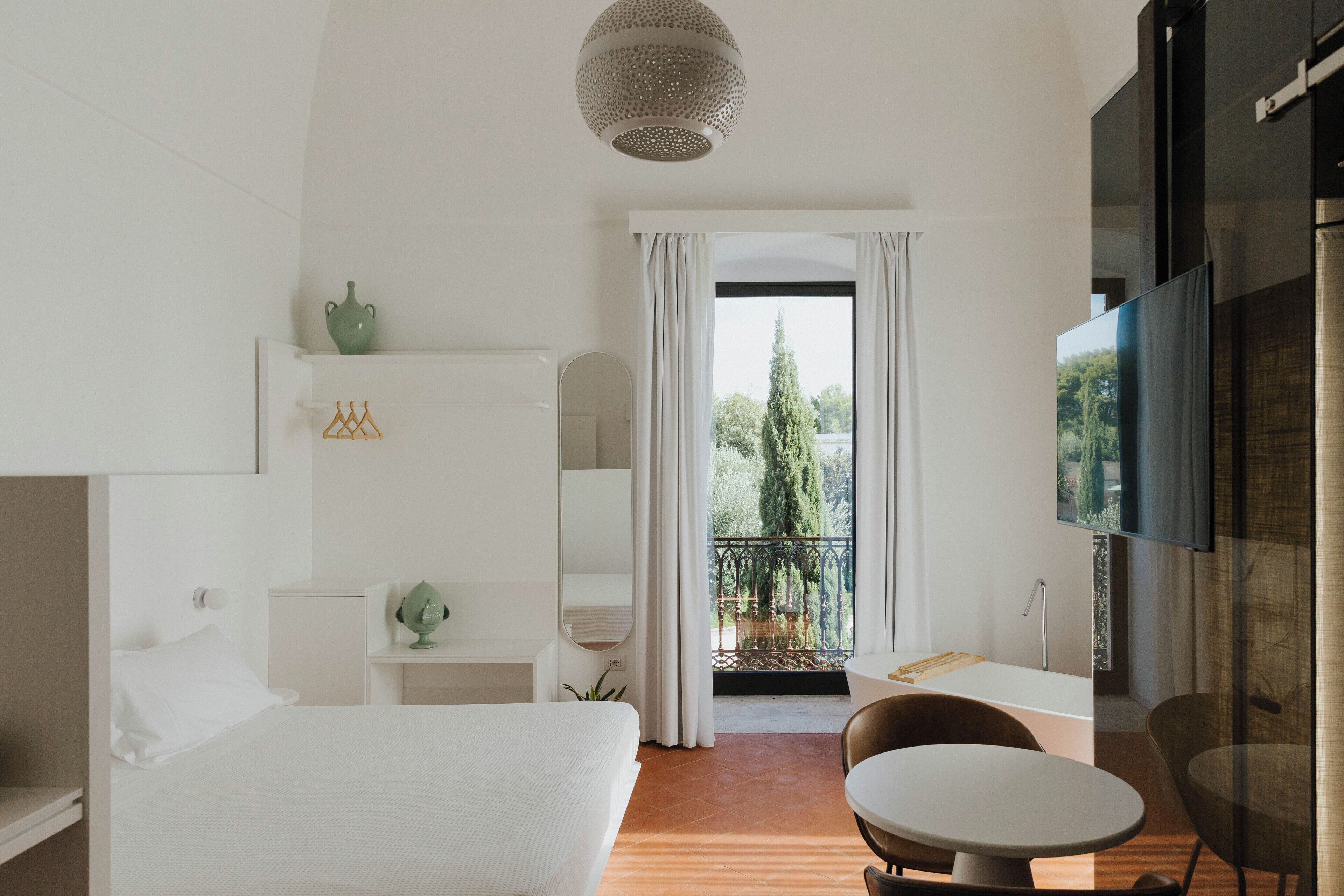 Une chambre d’hôtel moderne équipée d’un lit, d’une petite table, d’une chaise, d’une télévision et d’un balcon donnant sur la verdure.