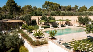 Zona de piscina con sillas de descanso y sombrillas, rodeada de vegetación y un muro de piedra.