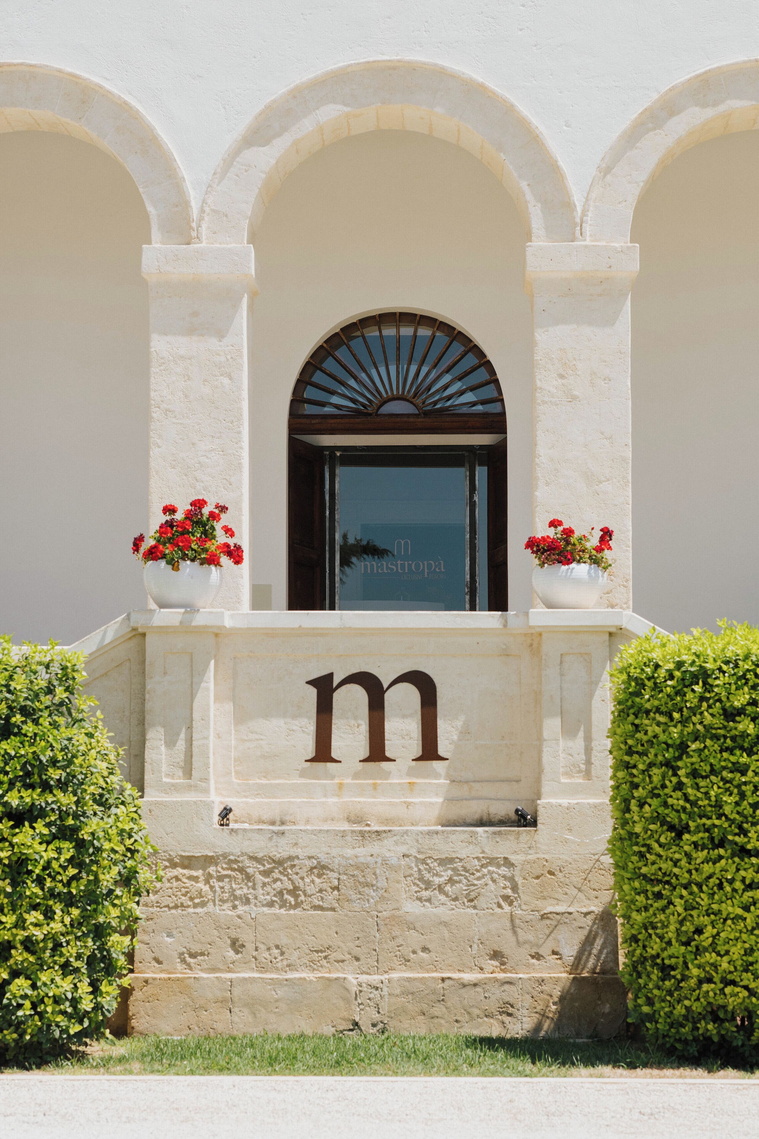 A building entrance with a large 'm' sign, a window with a sunburst design, and two flower pots on either side.