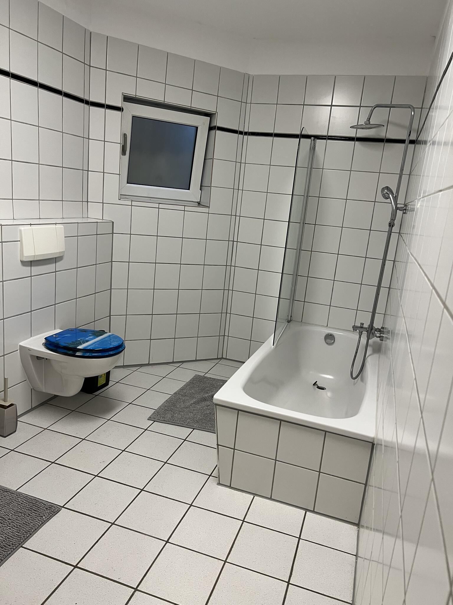 Kylpyhuone, jonka seinät on laatoitettu valkoisilla laatoilla, ja jossa on sinisellä istuimella varustettu wc, kylpyamme ja suihku.