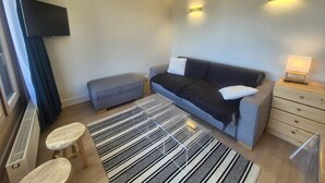 Ett modernt hotellrum med en grå soffa, en liten pall, ett träbord vid sängen med en lampa och ett stort fönster med gardiner.