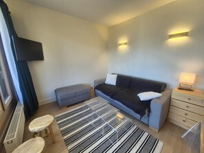 A modern hotel room with a grey sofa, a small ottoman, a wooden nightstand with a lamp, and a large window with curtains. - 100m from resort center, 2nd floor, view mountain, balcony, tv, ski locker, 40m², Tignes (Tignes)