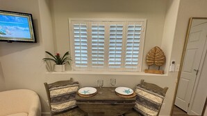 A dining area with a wooden table, two chairs, a TV mounted on the wall, a window with blinds, a potted plant, and a decorative shell on the wall.