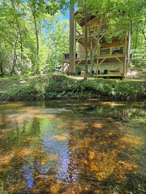 A wooden cabin with a deck and a river in front. - Creekside treehouse. with hot tub. (Piedmont)