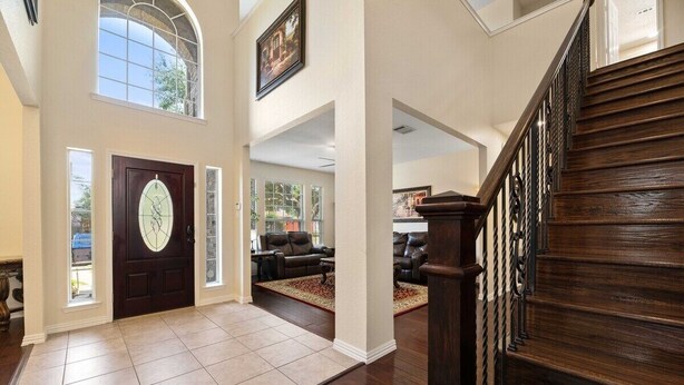 A spacious foyer with a dark wooden staircase, a large door with a decorative glass panel, and a view into a living area with leather sofas. - Secluded Luxe Home | Private Pool & Grill (Allen)