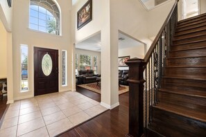 A spacious foyer with a dark wooden staircase, a large door with a decorative glass panel, and a view into a living area with leather sofas. - Secluded Luxe Home | Private Pool & Grill (Allen)