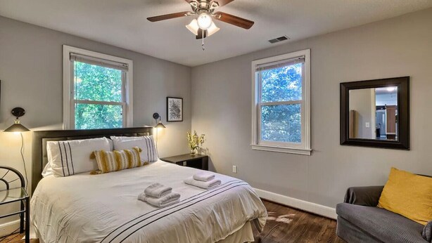 Quarto com cama, ventilador de teto, duas janelas, mesa de cabeceira, cadeira e espelho.