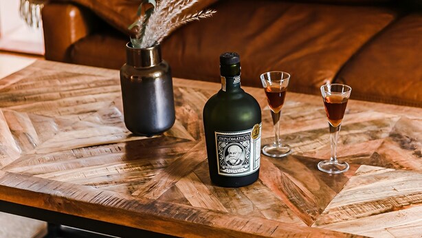 Een houten bijzettafeltje met een fles en twee glaasjes amberkleurige vloeistof, een vaas met gedroogde planten, en een bruine leren bank op de achtergrond.