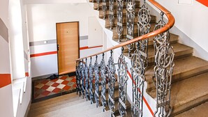Een trap met sierlijk metalen hekwerk en een houten leuning, die leidt naar een kamer met een geruit vloertapijt en een deur.