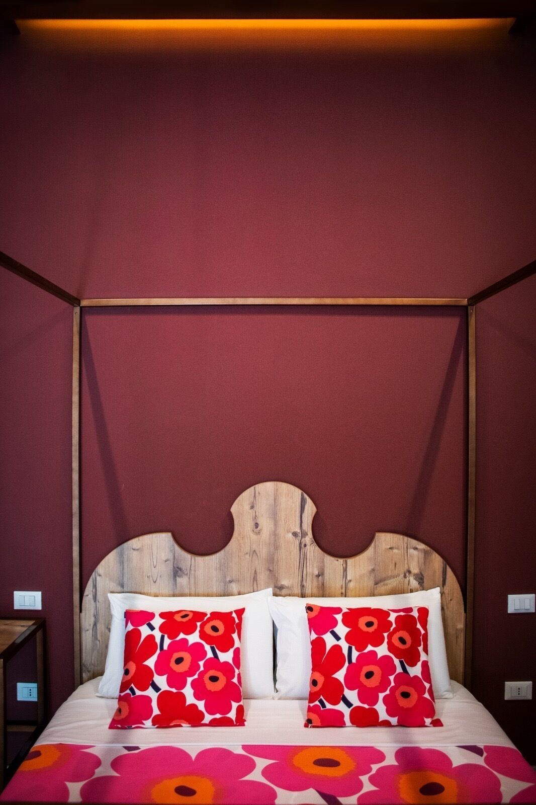 Una cama con cabecera de madera, dos almohadas rojas y blancas, y una colcha con estampado floral.