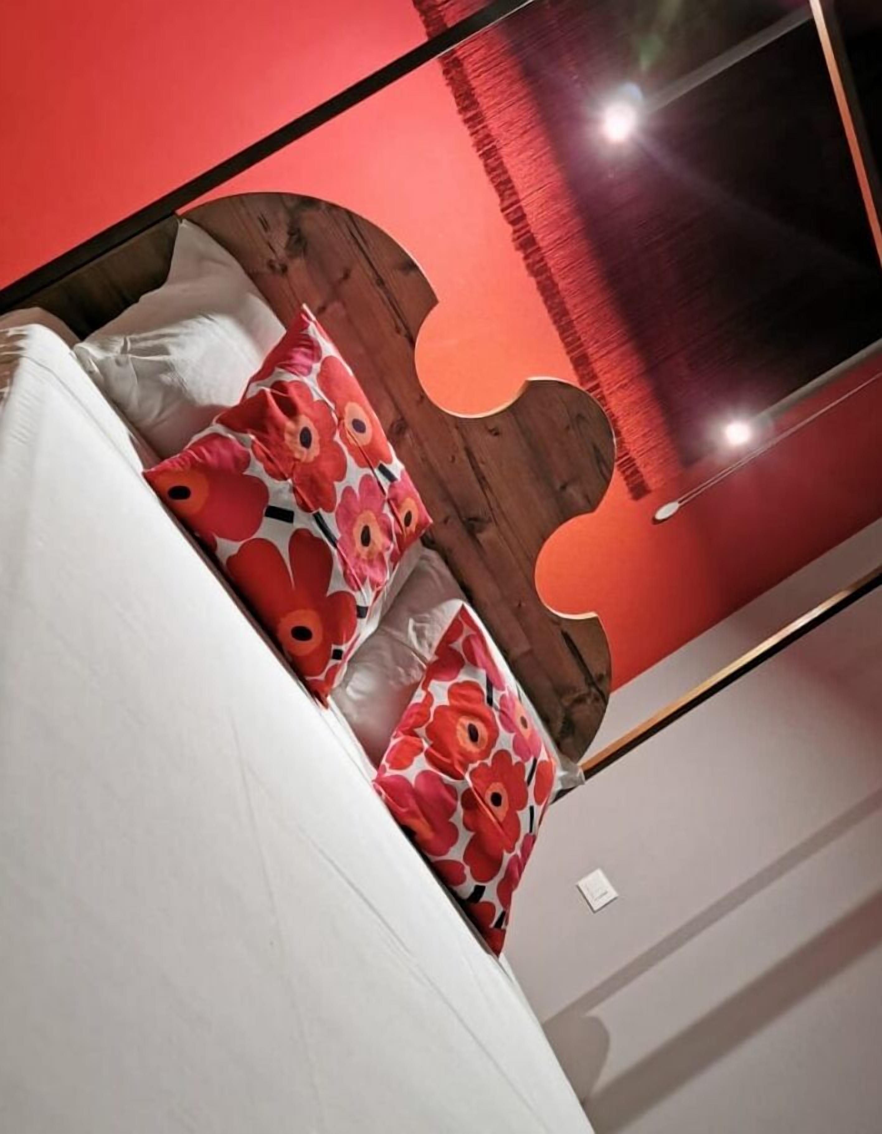 A modern bedroom with a white bed, red floral pillows, and a wooden headboard.