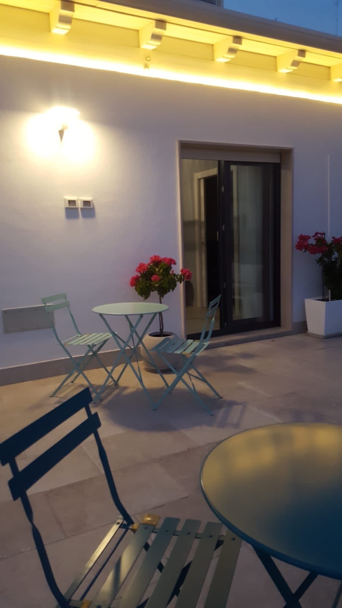 An outdoor patio area with a round table and two chairs, a potted plant with red flowers, and a wall-mounted light fixture.