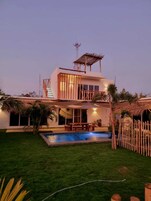 Una casa de dos pisos con piscina, un porche cubierto y una estructura de techo de paja.