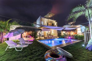 A modern house with a swimming pool, lounge chairs, and a thatched roof structure.
