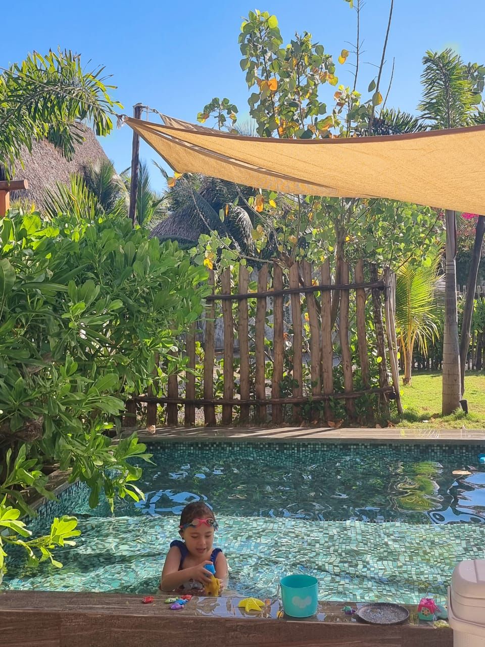A child playing in a pool surrounded by lush greenery and a wooden fence.