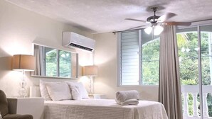 A bedroom with a bed, a ceiling fan, an air conditioning unit, a window with curtains, and a view of greenery outside. - #26 Beach Apartment 2br, 3ba w/ jet tub (Isabela)