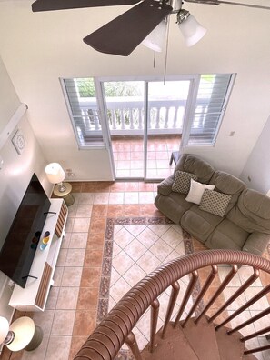 A living room with a sofa, a TV, a staircase, and a balcony with a railing. - #26 Beach Apartment 2br, 3ba w/ jet tub (Isabela)