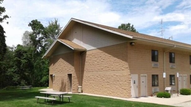 A building with a sloped roof, a flat entrance, and a small parking area with a picnic table. - Oakridge Motel (Newaygo)