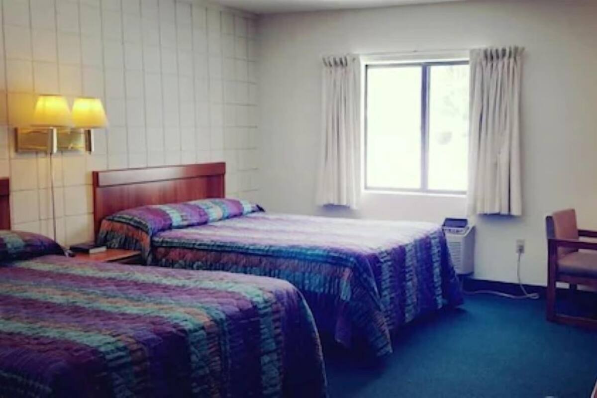 A hotel room with two beds, a desk, a chair, a window with curtains, and a wall-mounted lamp.