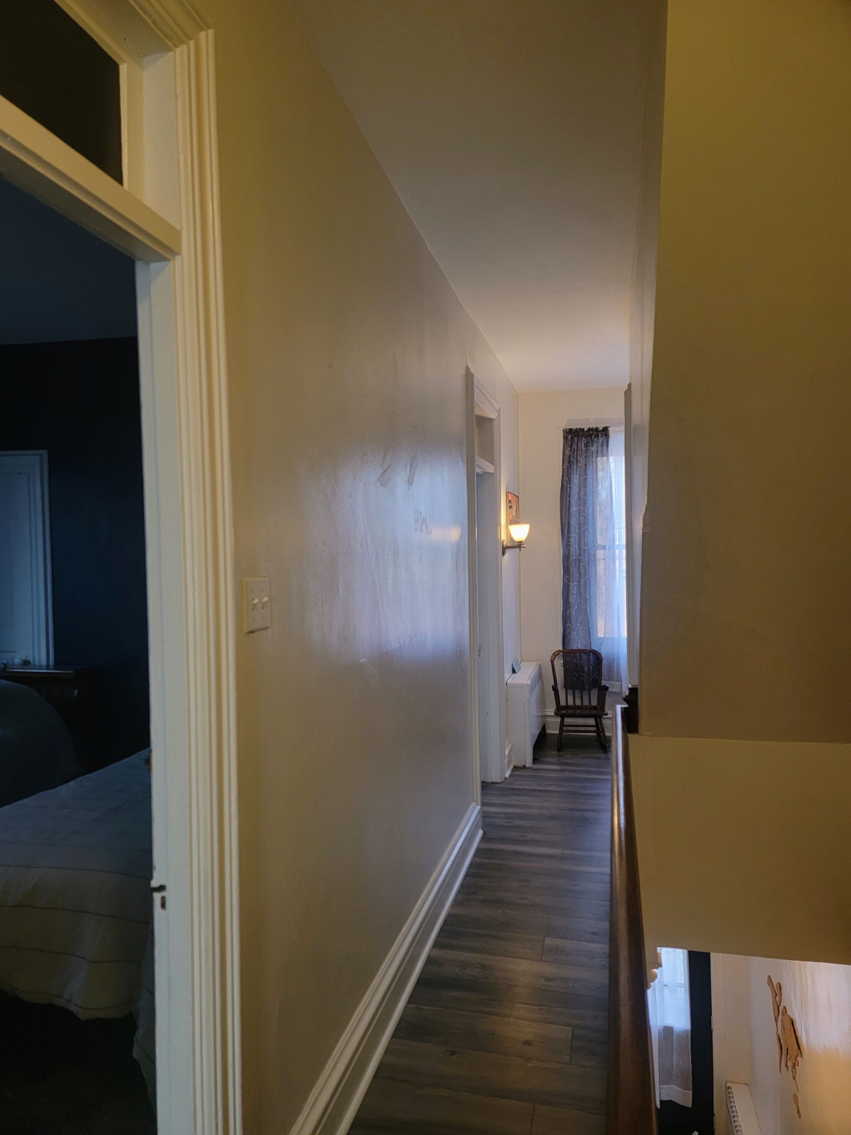 Un couloir doté d’un plancher en bois, de murs blancs et d’une porte donnant sur une pièce où se trouvent une chaise et une lampe.