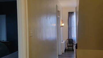Un couloir doté d’un plancher en bois, de murs blancs et d’une porte donnant sur une pièce où se trouvent une chaise et une lampe.